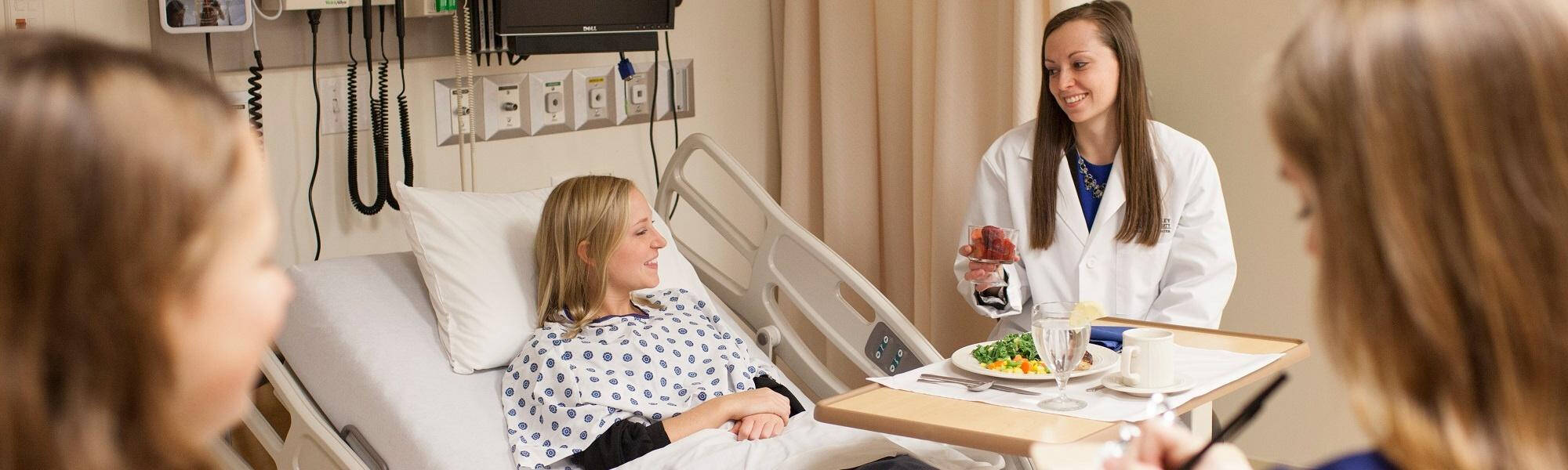 Clinical Dietetics students, one in a white coat and one in a hospital bed, complete a simulation exercise as two other students observe.